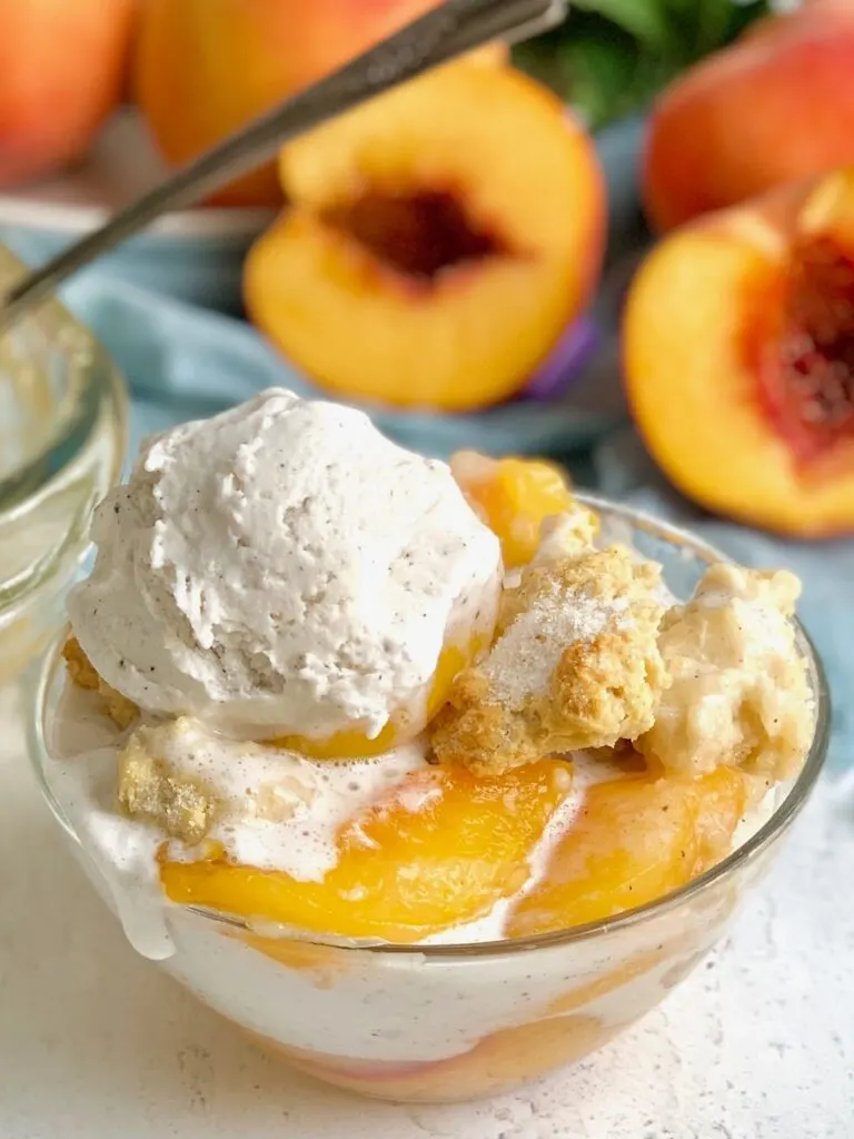 A close up of a dessert bowl filled with sugar peaches, golden brown cobbler topping and a scoop of melty vanilla ice cream. 