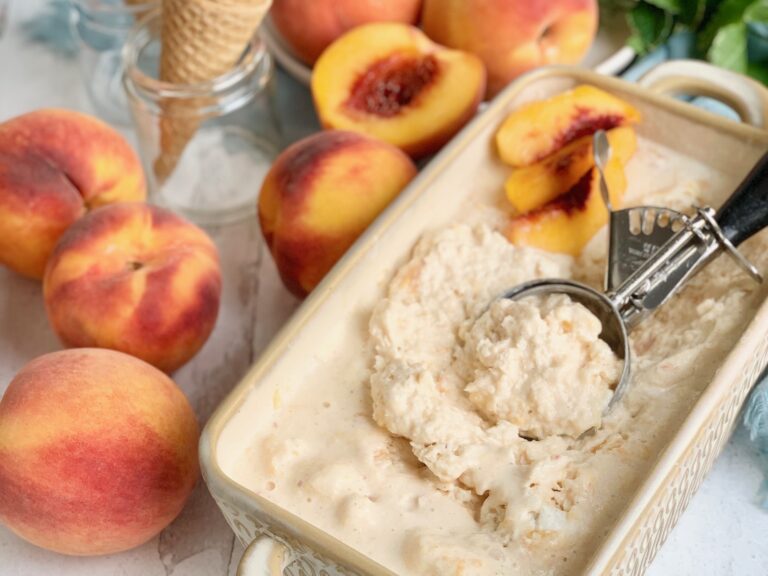 A bread loaf pan filled with peach flavored ice cream. An ice cream scoop is scooping some out. Nearby are several fresh peaches and an ice cream cone.