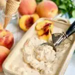A bread loaf pan filled with frozen peach colored ice cream next to several fresh peaches. An ice cream scoop is in the ice cream.