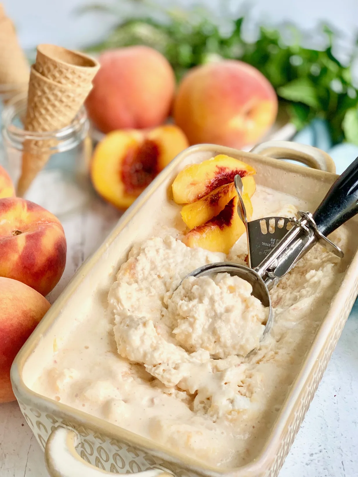 A bread loaf pan filled with frozen peach colored ice cream next to several fresh peaches. An ice cream scoop is in the ice cream.