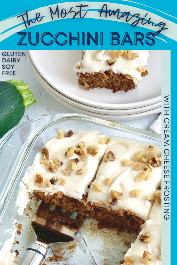 A glass pan holding rectangle golden colored bars with a creamy cream cheese frosting and chopped walnuts over the top. A serving spatula is getting one piece out next to a plates and a whole zucchini.