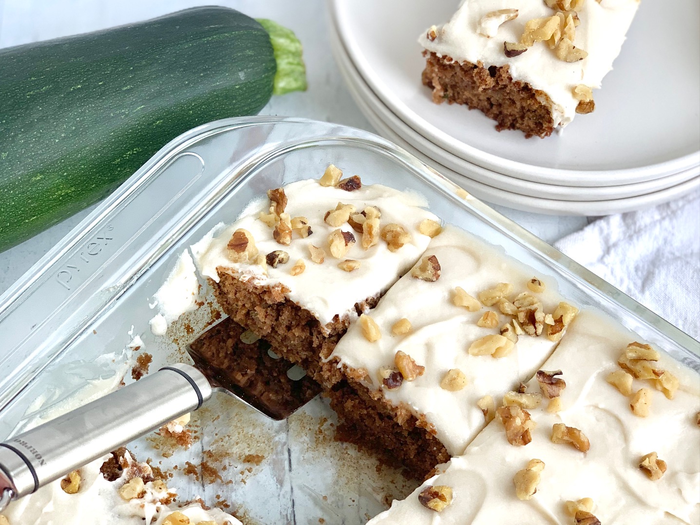 A glass baking pan filled with a golden colored cake and cream cheese frosting on top with scattered chopped walnuts next to a whole zucchini and white serving plates.