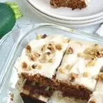 A glass pan holding rectangle golden colored bars with a creamy cream cheese frosting and chopped walnuts over the top. A serving spatula is getting one piece out.