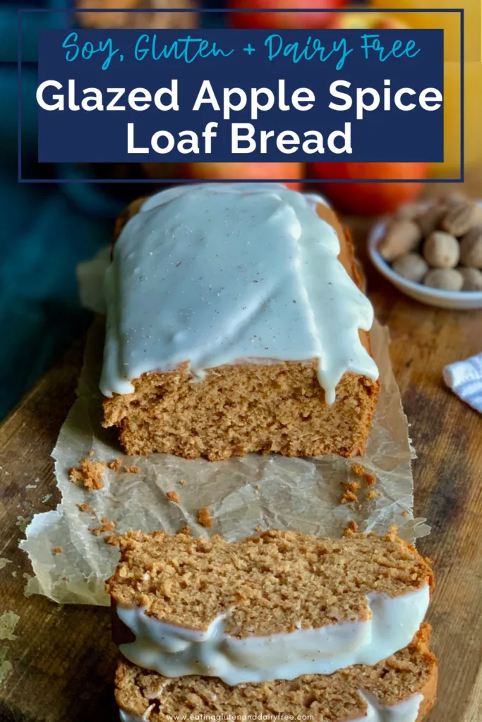 A golden colored loaf of bread with a white glaze on top cut into large bakery style slices next to fresh nutmeg, large juicy apples, and a glass of apple cider.