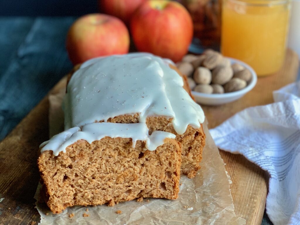 A cutting board with golden colored apple bread with a thick, white glaze over the top next to 3 red apples and a jar filled with apple cider.