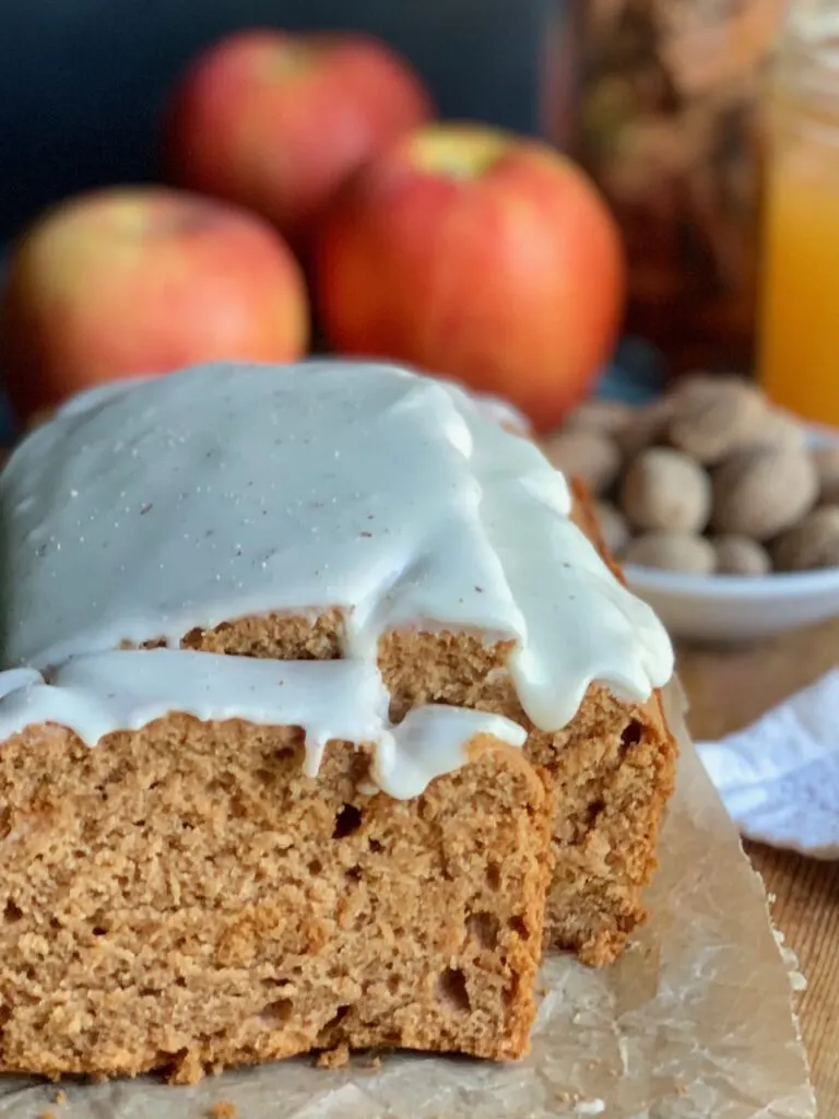 A golden colored loaf of bread cut into slices with a white glaze on top next to fresh nutmeg, large juicy apples, and a glass of apple cider.