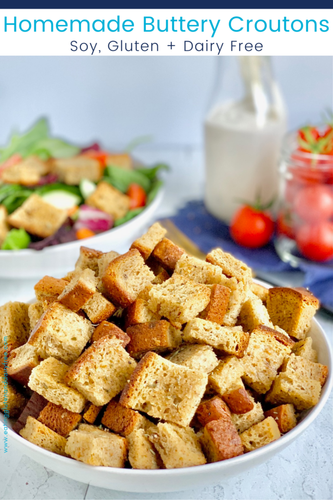 A large bowl with cubed, crispy, croutons in it with a salad in the background and mini grape tomatoes.