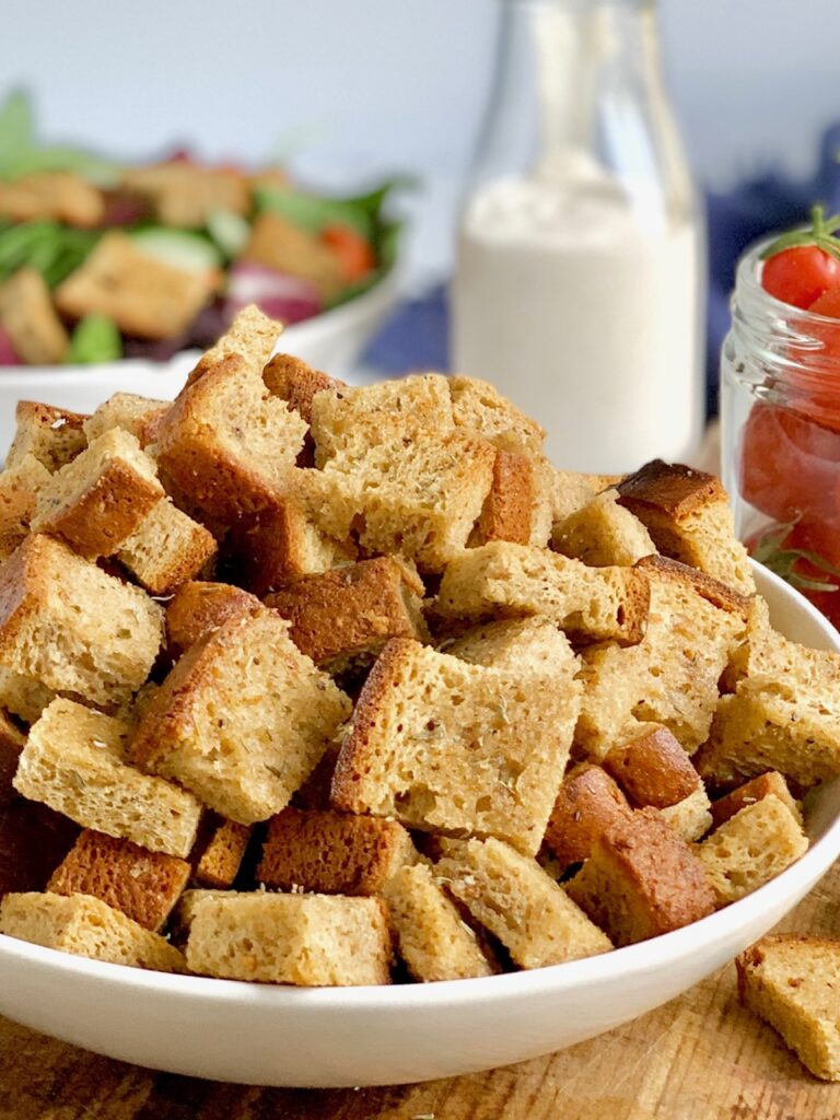 A large bowl with cubed, crispy, croutons in it next to a jar of grape tomatoes.