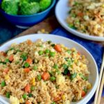 A large white plate piled with golden brown rice with small cubed carrots, peas, corn, and green beans next to chopsticks and a bowl of broccoli.