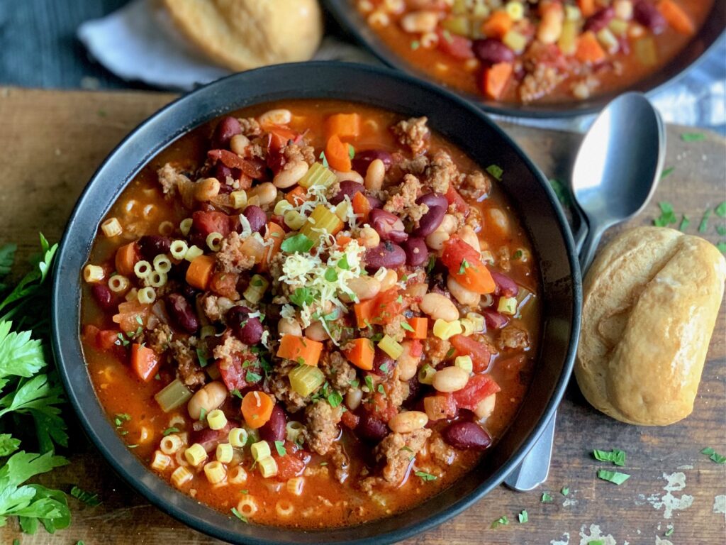 Two black bowls filled with ground sausage, celery, carrots, onions, two different kinds of beans, in a fire roasted broth next to dinner rolls and spoons.