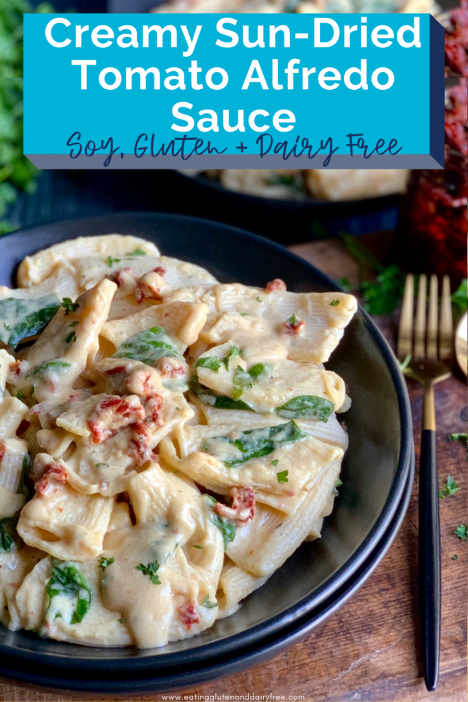 A large black bowl with small pasta noodles covered in a creamy sauce with red sun-dried tomatoes and spinach in the sauce next to a gold fork and spoon. With a small glass jar of sun-dried tomatoes in it.