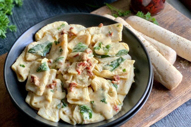 A large black bowl filled with tender pasts noodles coated in a white alfredo sauce with lots of spinach and chopped sun dried tomatoes on a cutting board next to a few baked garlic breadsticks.