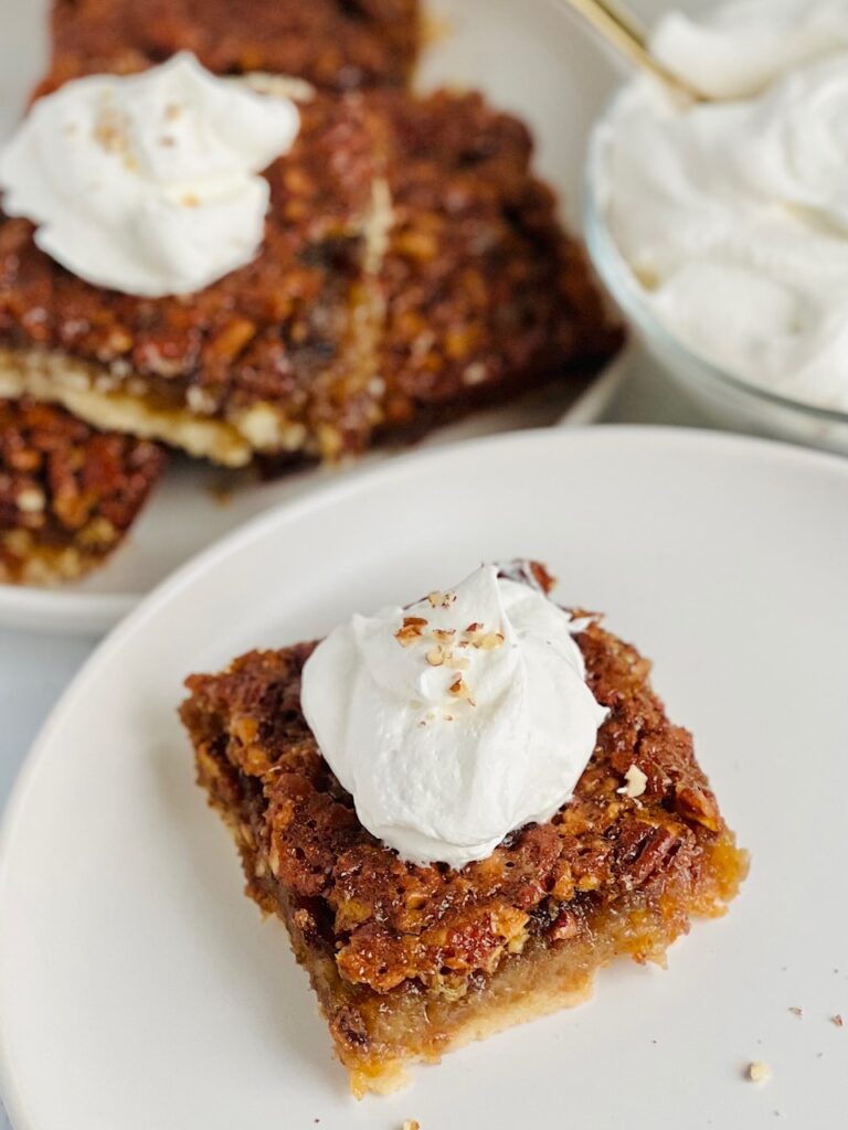 A golden brown bar cut into a square with whipped topping on it next to a plate of several bars.