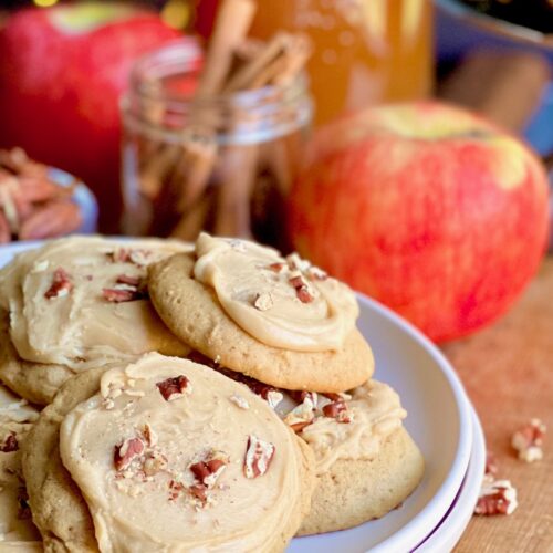 A white plate full of small golden colored cookies with a brown frosting on top and chopped nuts next to a jar of cinnamon sticks and a few large red apples.