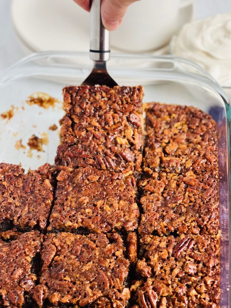 A glass baking pan with a pecan dessert. the pan is cut into large squares and a serving spatula is lifting a sqaure out. 