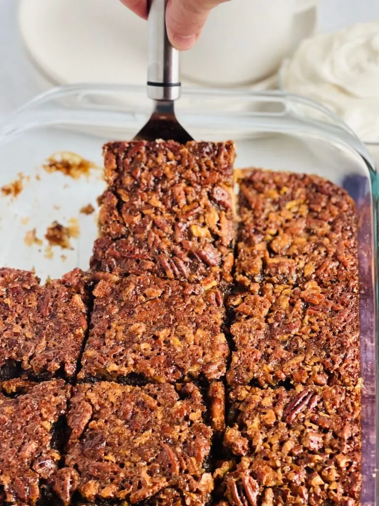 A glass 9x13-inch pan with a golden pecan topping cut into 12 large bars and a serving spatula lifting one bar out of the pan.