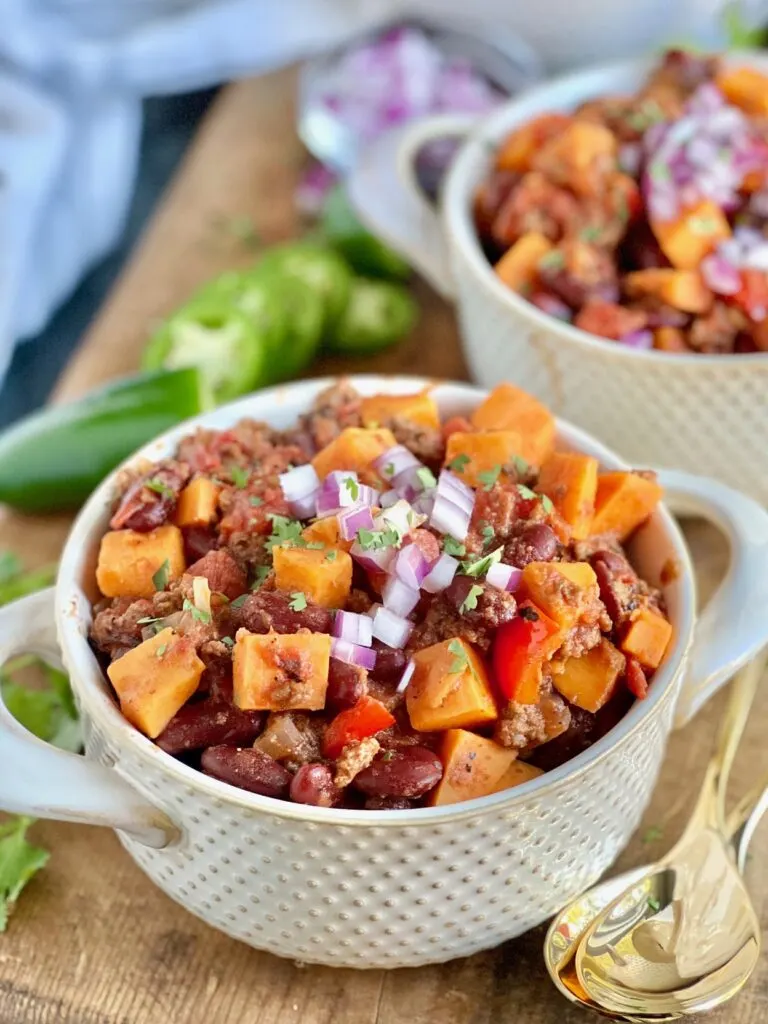 A mug of tender sweet potato chunks, beans, ground beef. and diced red bell peppers,  topped with diced red onions.