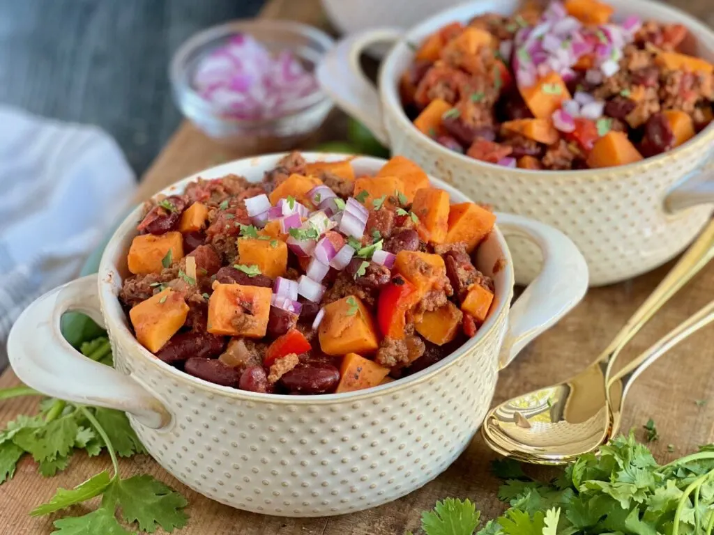 A mug of tender sweet potato chunks, beans, ground beef. and diced red bell peppers,  topped with diced red onions on a dark wood cutting board with fresh cilantro.