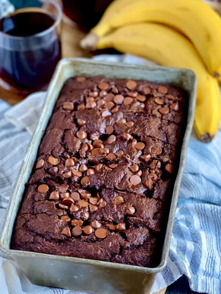 A metal loaf pan with a dark brown bread in it and chocolate chips on top. You can also see 2 bananas next to the loaf pan