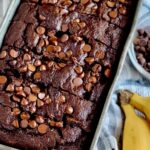 A metal loaf pan with a dark brown bread in it and chocolate chips on top. You can also see 2 bananas next to the loaf pan.