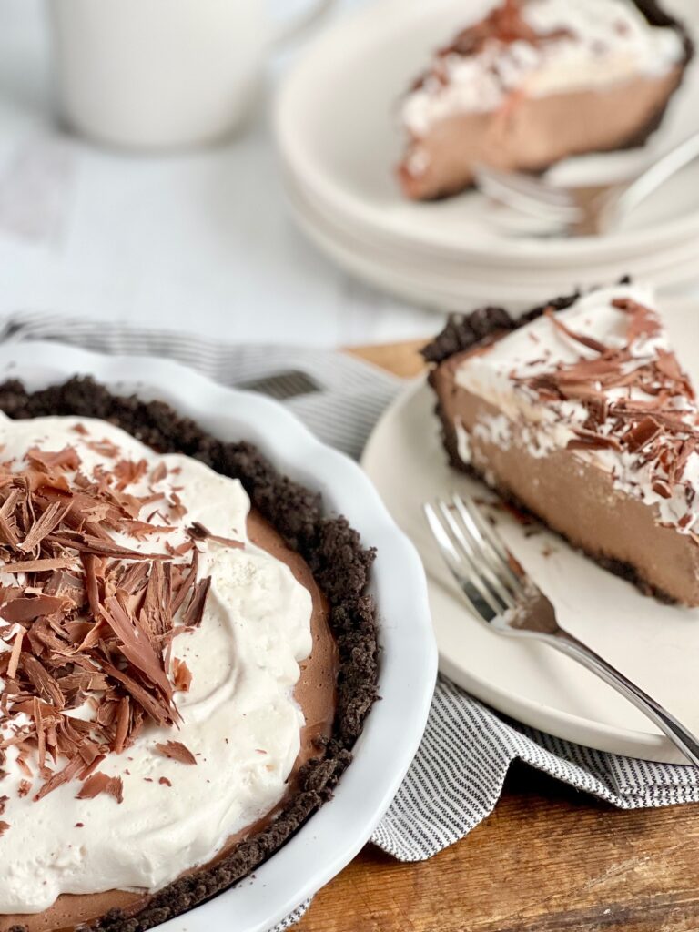 A white pie plate with a dark brown crust, a brown chocolate pudding, and topped with a white whipped topping. Last on top of that is small pieces of hard chocolate. Nearby  are two plates of pie on them with a fork.