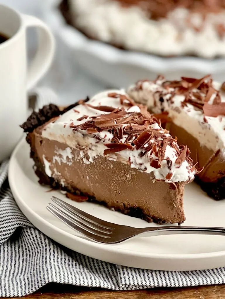 Two slices of pie on a white plate. The pie crust is dark brown and make out of oreo cookies and butter. The middle of the pie is a thick chocolate pudding, and the top of the pie is frozen whipped topping with chocolate curls.