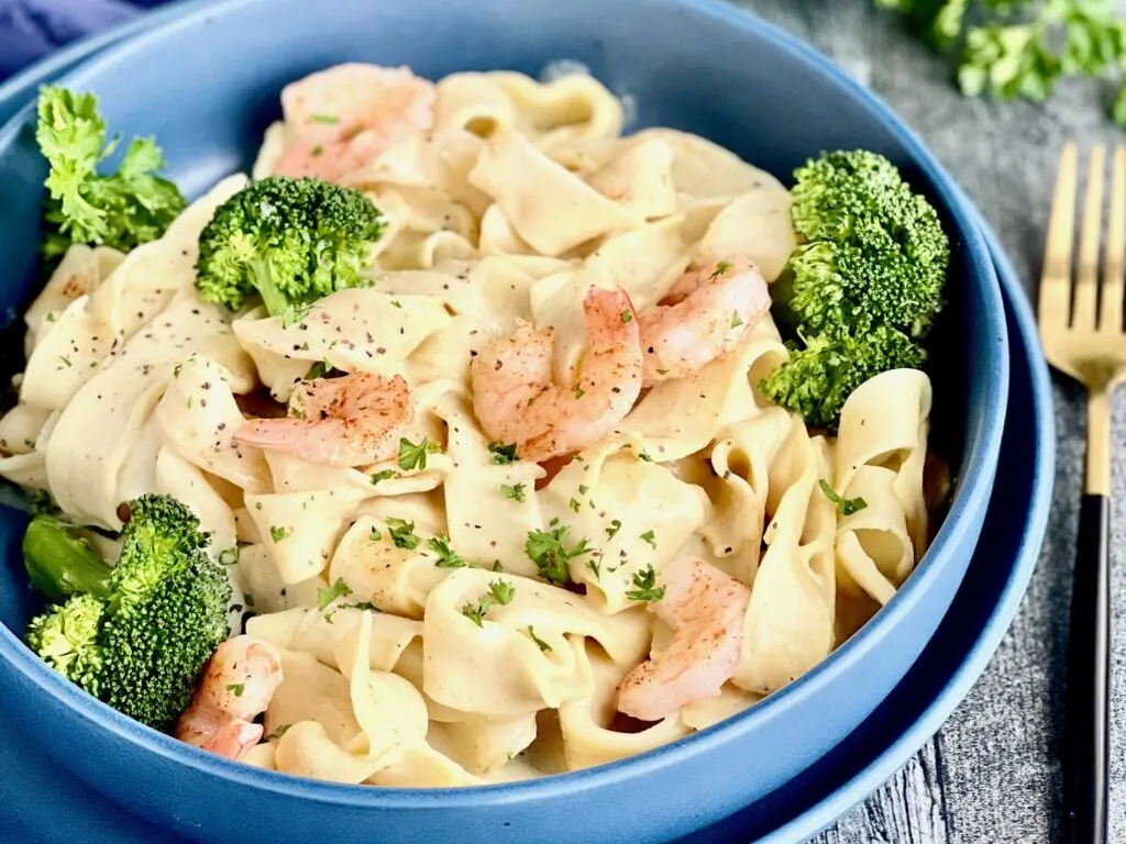 A blue blue filled with white thick pasta noodles and a creamy looking white sauce with saut&eacute;ed shrimp and steamed green broccoli. There are fresh chopped pieces of green parsley on top,