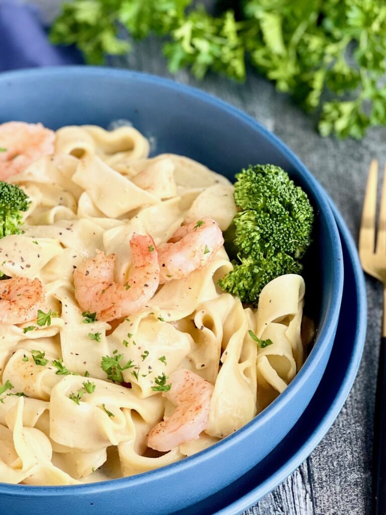 A blue blue filled with white thick pasta noodles and a creamy looking white sauce with sautéed shrimp and steamed green broccoli. There are fresh chopped pieces of green parsley on top with a gold colored fork nearby.