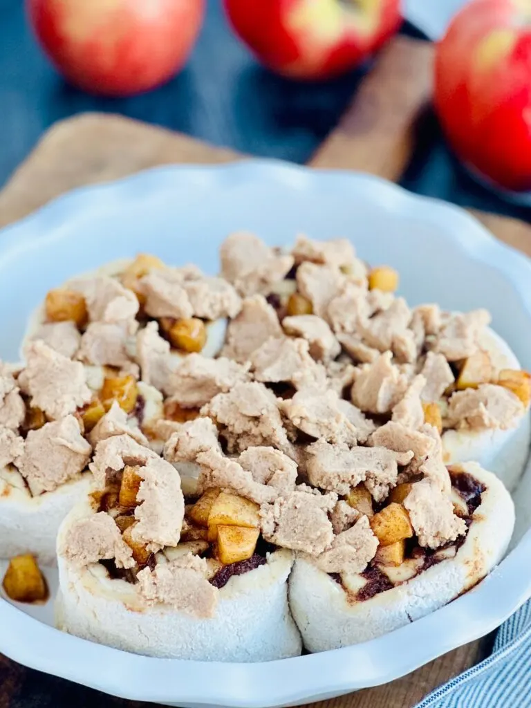 A white pie plate with 7 round dough ball cinnamon rolls with apple chunks on top. And a thick mixture in litttle pieces like a crumble over that.