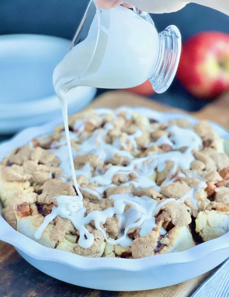 A white pie plate with 7 round dough ball cinnamon rolls with a crumble topping and someone drizzling white icing over the top.