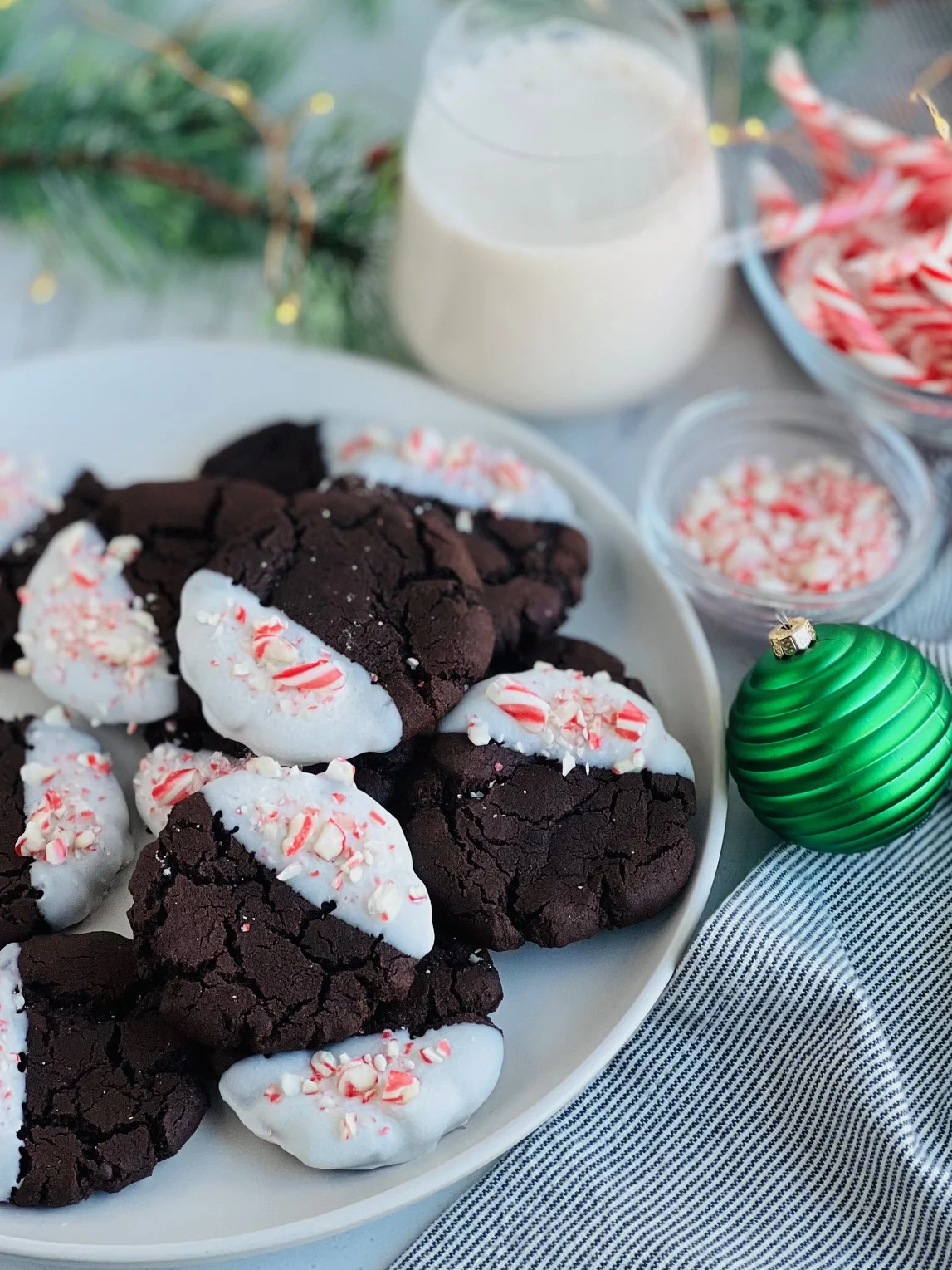 Dark chocolate colored round cookies with half of the cookie coated with a white glaze and chopped candy canes on a white plate next to a cup of almond milk.