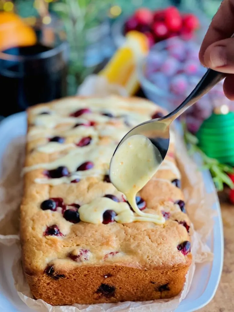 A white bread dish with a cream colored rectangle loaf  with red circle cranberries popping out of the bread. A light orange colored glaze is being drizzled over the top with a spoon.