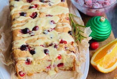 A white bread dish with a cream colored rectangle loaf with red circle cranberries popping out of the bread. A light orange colored glaze is drizzled over the top.