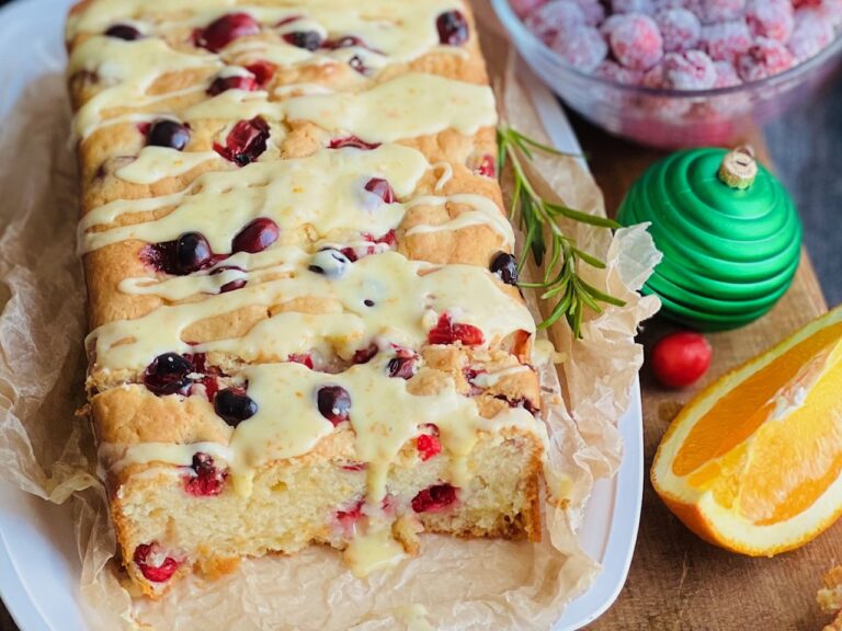 A white bread dish with a cream colored rectangle loaf with red circle cranberries popping out of the bread. A light orange colored glaze is drizzled over the top.