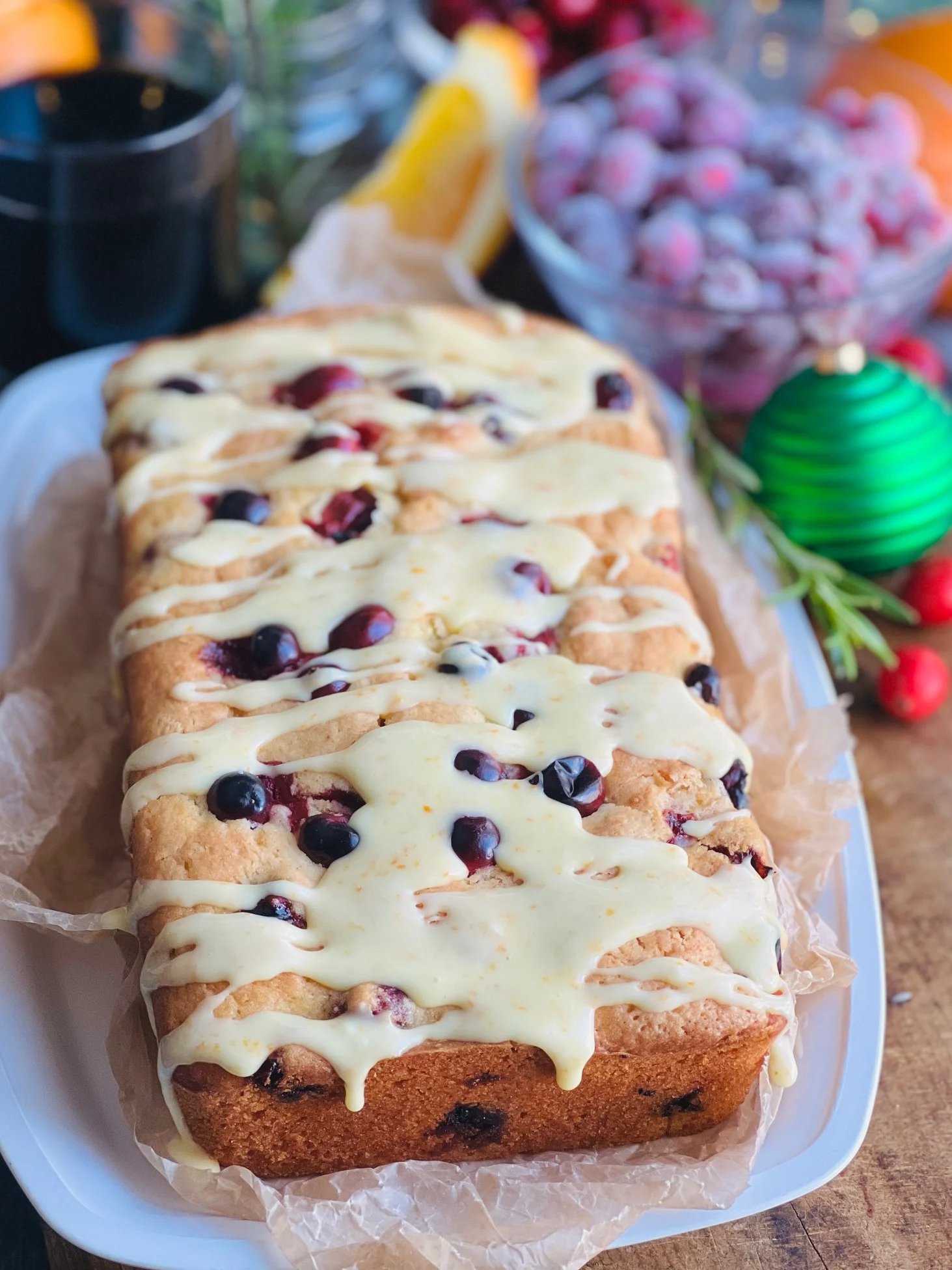 A white bread dish with a cream colored rectangle loaf with red circle cranberries popping out of the bread. A light orange colored glaze is drizzled over the top.