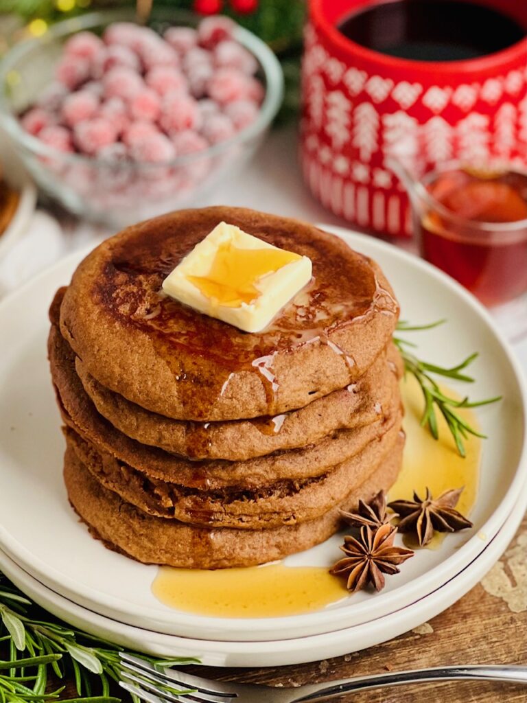A white plate filled with dark brown colored round pancakes with a large square of melty butter and syrup running down the top and forks nearby.
