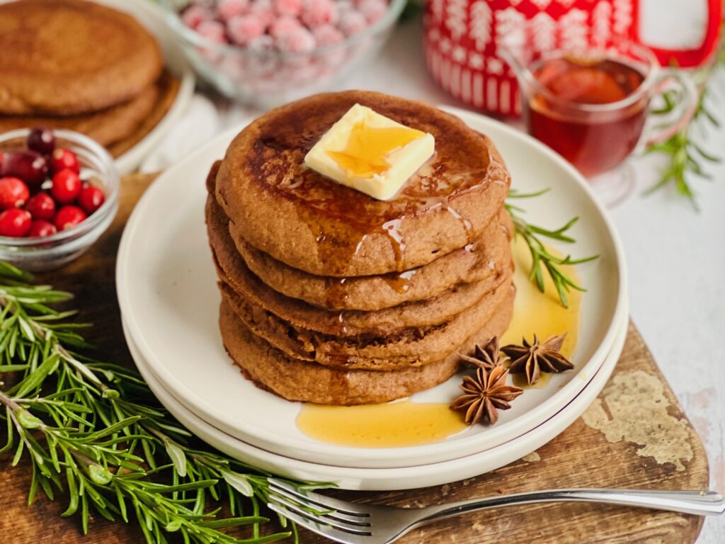 A white plate filled with dark brown colored round pancakes with a large square of melty butter and syrup running down the top and a small bowl of cranberries and syrup nearby.