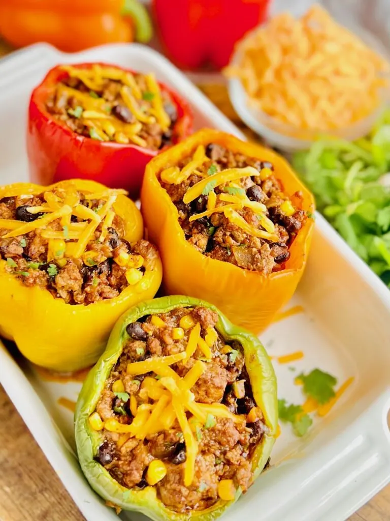 A white baking dish filled with 1yellow, 1 red, 1 green, and 1 orange  bell pepper with the tops cut off. Inside is a seasoned meat sauce with corn, black beans, beef, and shredded cheese. There is dollop of dairy free sour cream on each baked pepper.