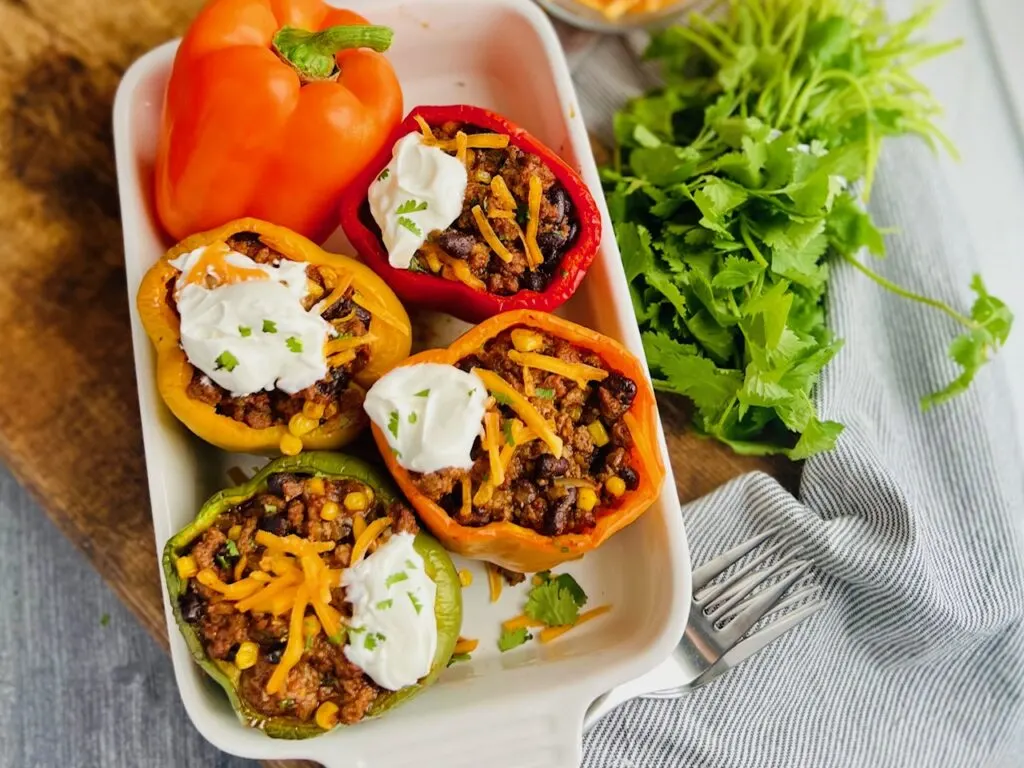 A white baking dish filled with 1yellow, 1 red, 1 green, and 1 orange  bell pepper with the tops cut off. Inside is a seasoned meat sauce with corn, black beans, beef, and shredded cheese. There is dollop of dairy free sour cream on each baked pepper.