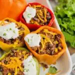 A white baking dish filled with 1yellow, 1 red, 1 green, and 1 orange bell pepper with the tops cut off. Inside is a seasoned meat sauce with corn, black beans, beef, and shredded cheese. There is dollop of dairy free sour cream on each baked pepper.
