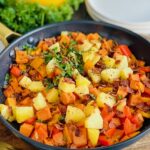 A black frying pan packed with sautéed sweet potatoes, diced apples, red and orange bell peppers, and topped with bits of bacon and Italian parsley next to white breakfast plates.