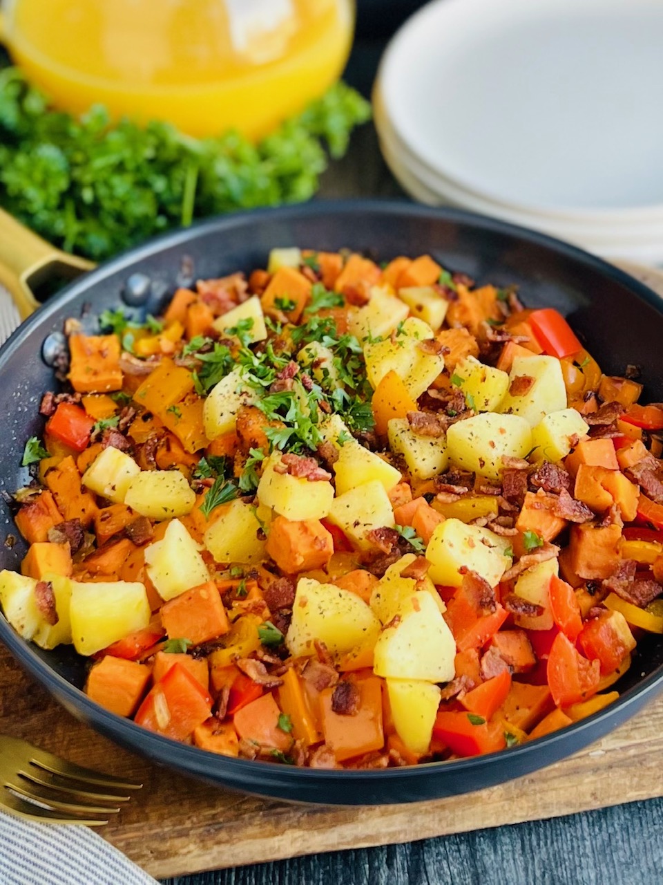 A black frying pan packed with sautéed sweet potatoes, diced apples, red and orange bell peppers, and topped with bits of bacon and Italian parsley next to white breakfast plates.