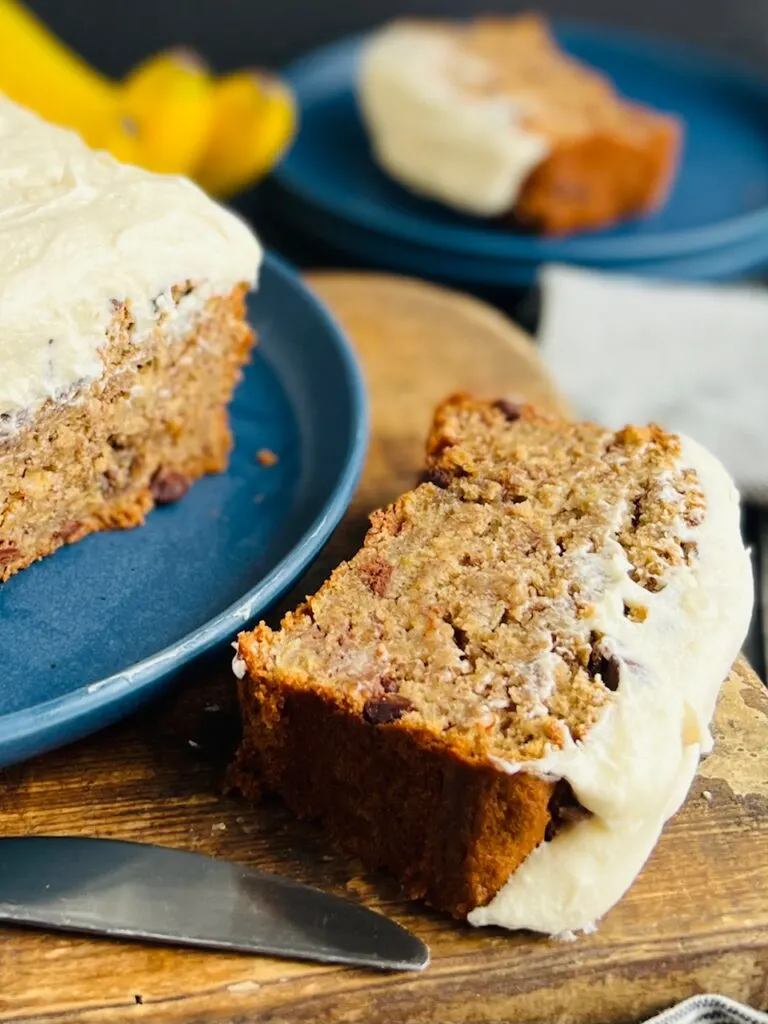 A blue round plate with a large loaf of banana bread with semisweet chocolate chips on it cut into slices. A large slice is near the plate with white and creamy frosting.