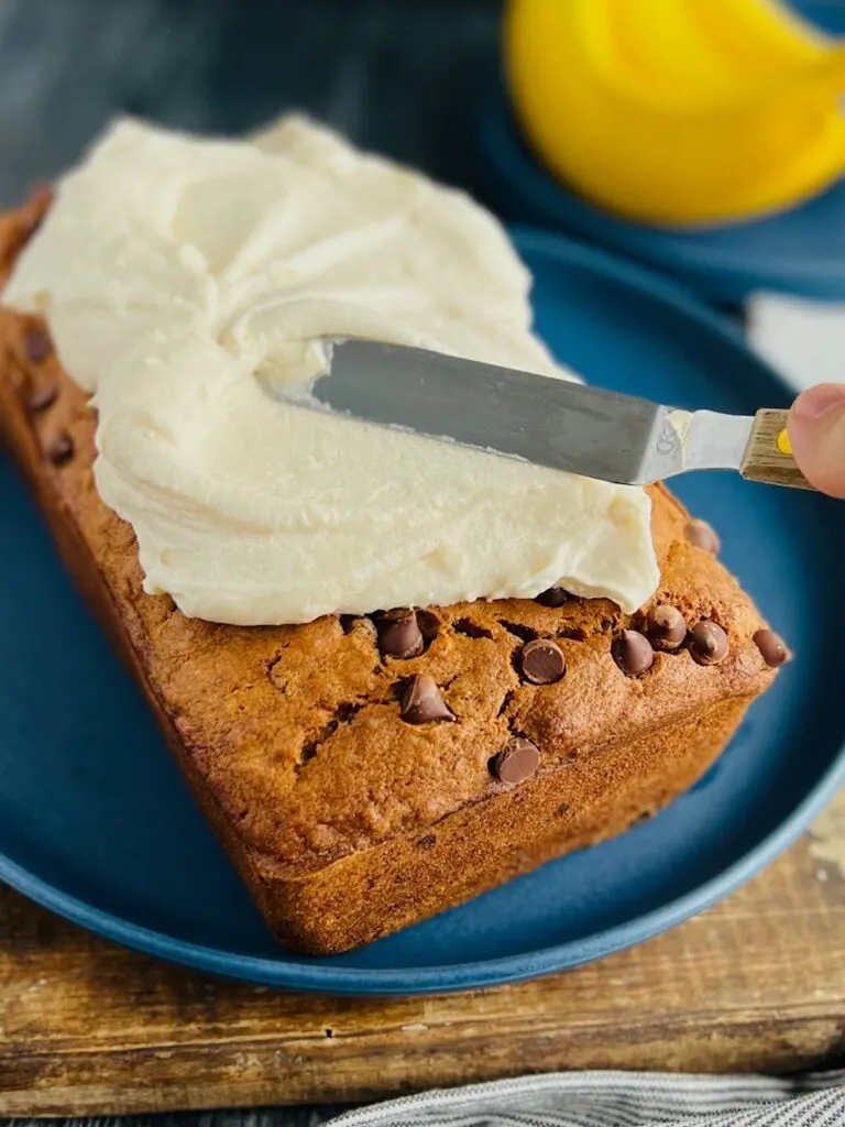 A blue round plate with a large loaf of banana bread with semisweet chocolate chips on it. White and creamy frosting is being put on the top with an angled spatula.