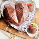 A chocolate heart sitting on a decorative plate with chocolate covered strawberries nearby.