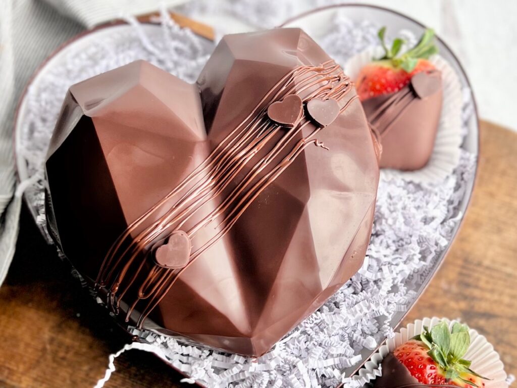 A chocolate heart sitting on a decorative plate with chocolate covered strawberries nearby. There is melted chocolate drizzled across the front.