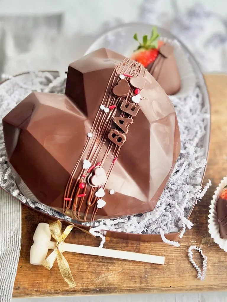 A chocolate heart sitting on a decorative plate with chocolate covered strawberries nearby. There is melted chocolate drizzled across the front with red and white sprinkles and the word BAE in chocolate.