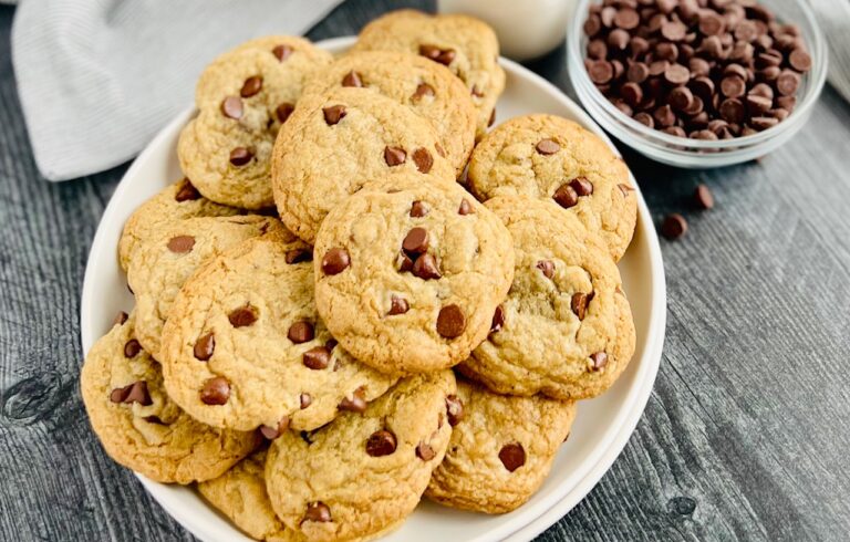 A round white plate with layers of chocolate chip cookies on it next to a small bowl of semisweet chocolate chips.