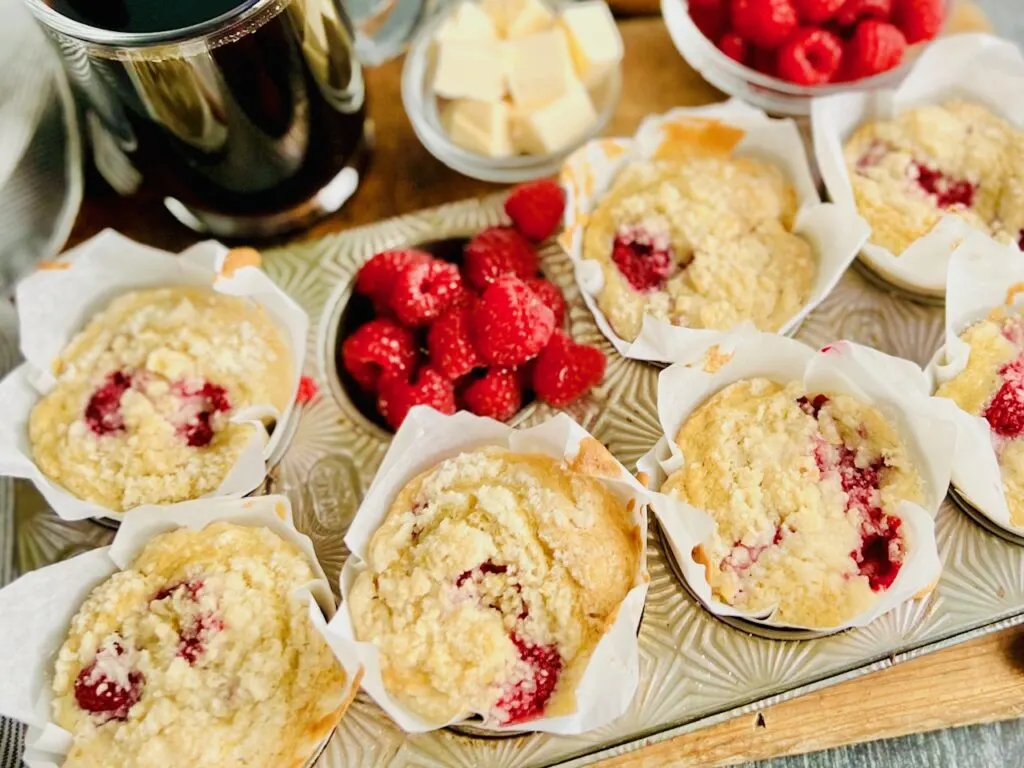 A silver muffin tin holding 7 light colored muffins with speckles of red raspberries popping through. One muffin cavity is filled with bright red raspberries. Next to the muffin tin is a mug of coffee and little cubes of cut up butter.