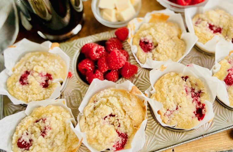 A silver muffin tin holding 7 light colored muffins with speckles of red raspberries popping through. One muffin cavity is filled with bright red raspberries. Next to the muffin tin is a mug of coffee and little cubes of cut up butter.